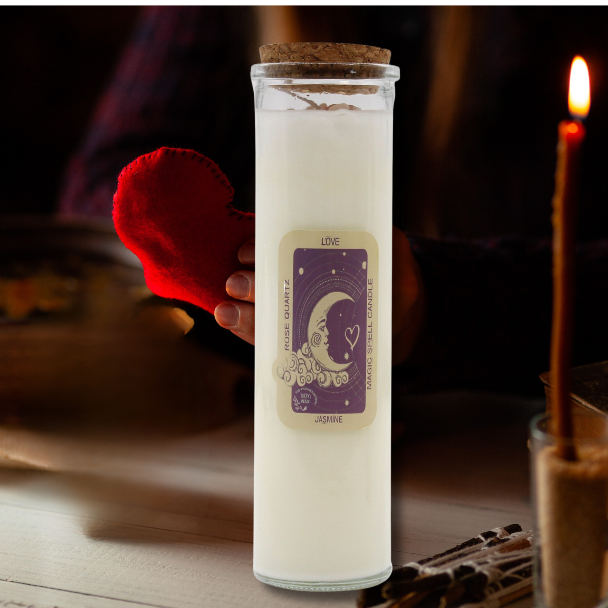 White candle in a glass jar with a label, held by a hand, on a wooden surface.